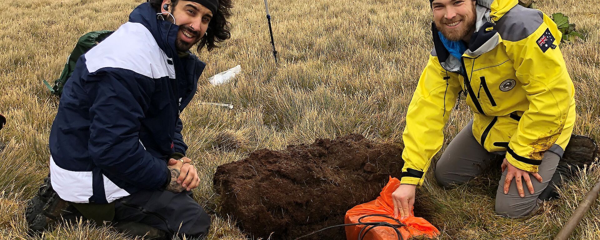 Two men sit next to a hole they have dug in to the grassy hillside.  One is lowering a seismometer in to the hole as both smile to camera.