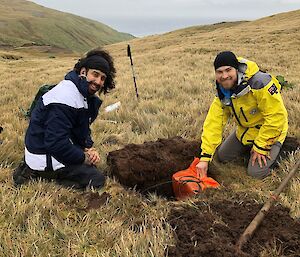 Two men sit next to a hole they have dug in to the grassy hillside.  One is lowering a seismometer in to the hole as both smile to camera.