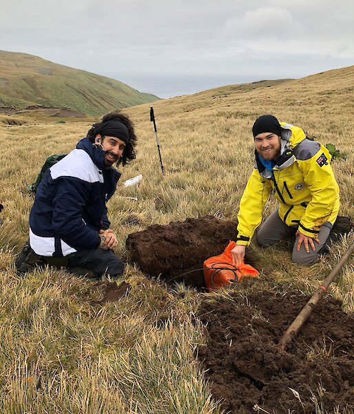 Two men sit next to a hole they have dug in to the grassy hillside.  One is lowering a seismometer in to the hole as both smile to camera.