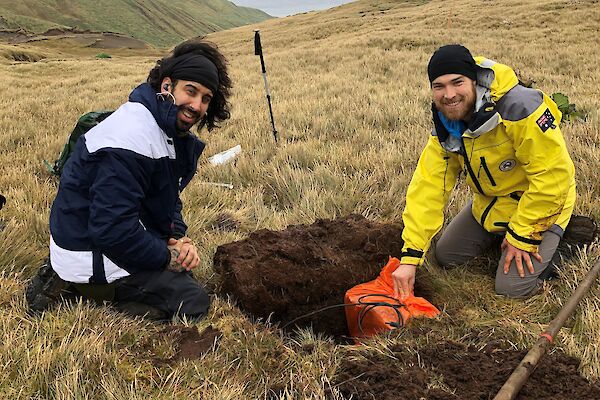 Two men sit next to a hole they have dug in to the grassy hillside. One is lowering a seismometer in to the hole as both smile to camera.
