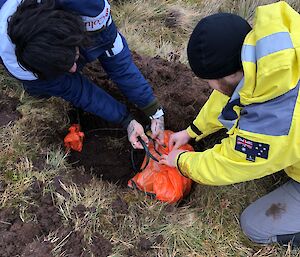 An overhead shot of two men dropping a seismometer into a hole dug in to the hillside