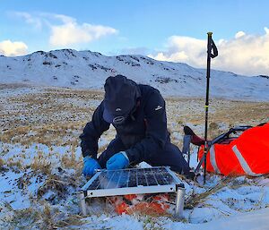 A man kneels in the snowy grass, snow topped mountains behind him,  He is fixing a screw in to a small solar panel.