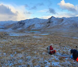 A beautiful snowy landscape of a tussocky plateau with snow topped mountain ridges in the distance.  A man can be seen working on a solar panel in the foreground.