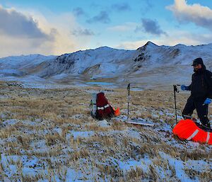 A beautiful snowy landscape of a tussocky plateau with snow topped mountain ridges in the distance.  A man stands in the foreground next to a recently installed solar panel.