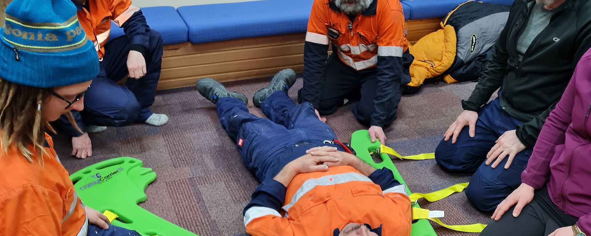 An expeditioner laying on the floor with a green split stretcher about to be slid under him by the five people kneeling around him.