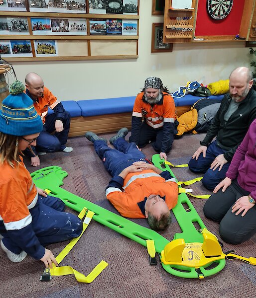 An expeditioner laying on the floor with a green split stretcher about to be slid under him by the five people kneeling around him.