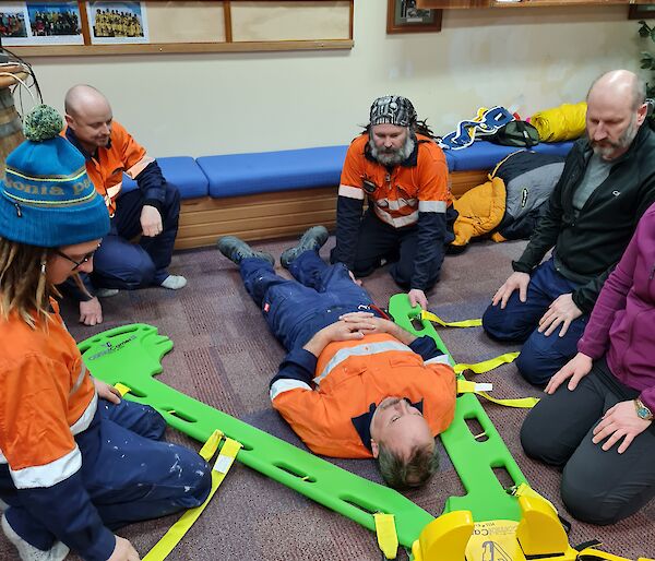 An expeditioner laying on the floor with a green split stretcher about to be slid under him by the five people kneeling around him.
