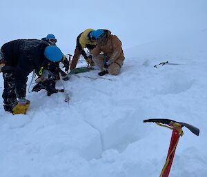 Four expeditioners in the snow, wearing hard hats with ropes, working together to create a snow anchor.  An ice picks sits in the snow in the foreground.