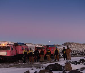 A group of people standing by 2 Haggs with a moon on the horizon.