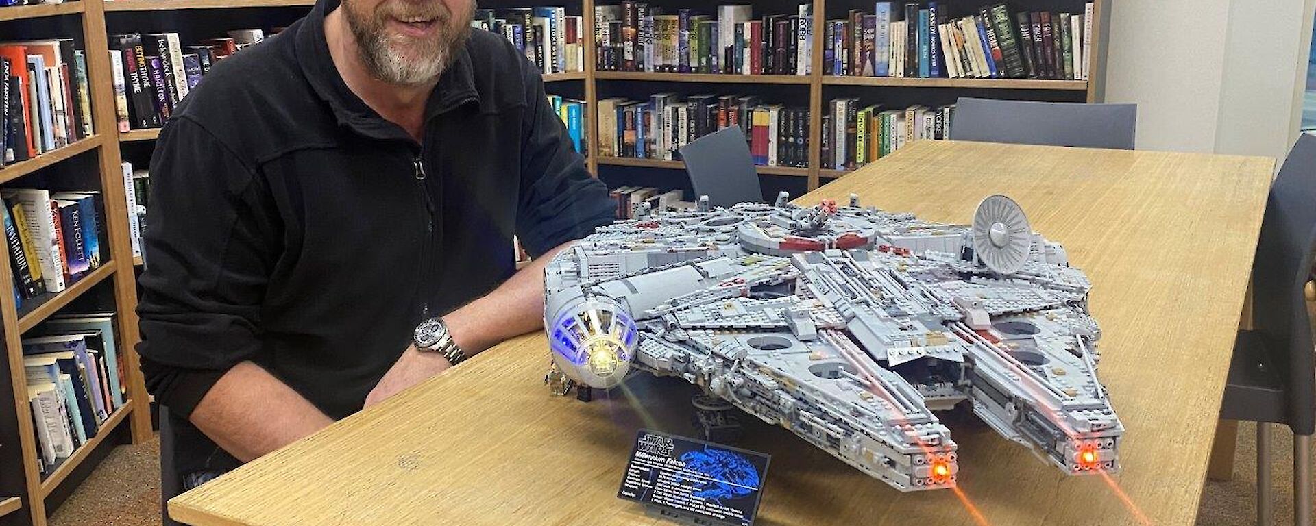 A man sits at a table, with bookshelves behind, smiling to camera.  A Lego Millennium Falcon model sits on the table, complete with lights and laser beams.