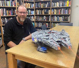 A man sits at a table, with bookshelves behind, smiling to camera.  A Lego Millennium Falcon model sits on the table, complete with lights and laser beams.