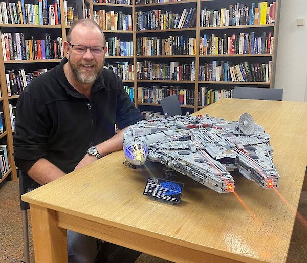 A man sits at a table, with bookshelves behind, smiling to camera.  A Lego Millennium Falcon model sits on the table, complete with lights and laser beams.
