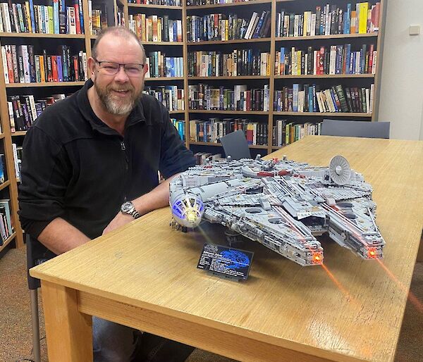 A man sits at a table, with bookshelves behind, smiling to camera. A Lego Millennium Falcon model sits on the table, complete with lights and laser beams.