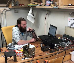 A man sits at a desk covered in electronic equipement speaking in to a small microphone