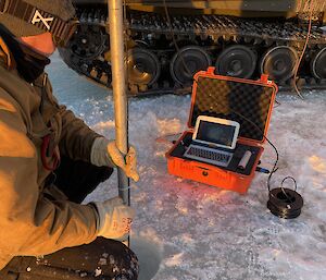 Expeditioner sititng on the sea ice, operating a long pole with underwater camera attached, looking at screen to locate tide gauge