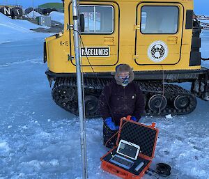 Expeditioner on sea ice with laptop downloading data from tide gauge below.  A yellow Hagglund in the background.
