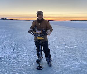 Expeditioner standing on the sea ice holding the petrol Jiffy drill with sunset in background