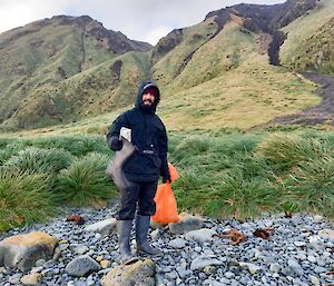 A man in all weather gear, stands on a pebbled beach with green tussocks and hills behind him.  He is holding an orange bin bag in one hand and a washed up gumboot in the other.