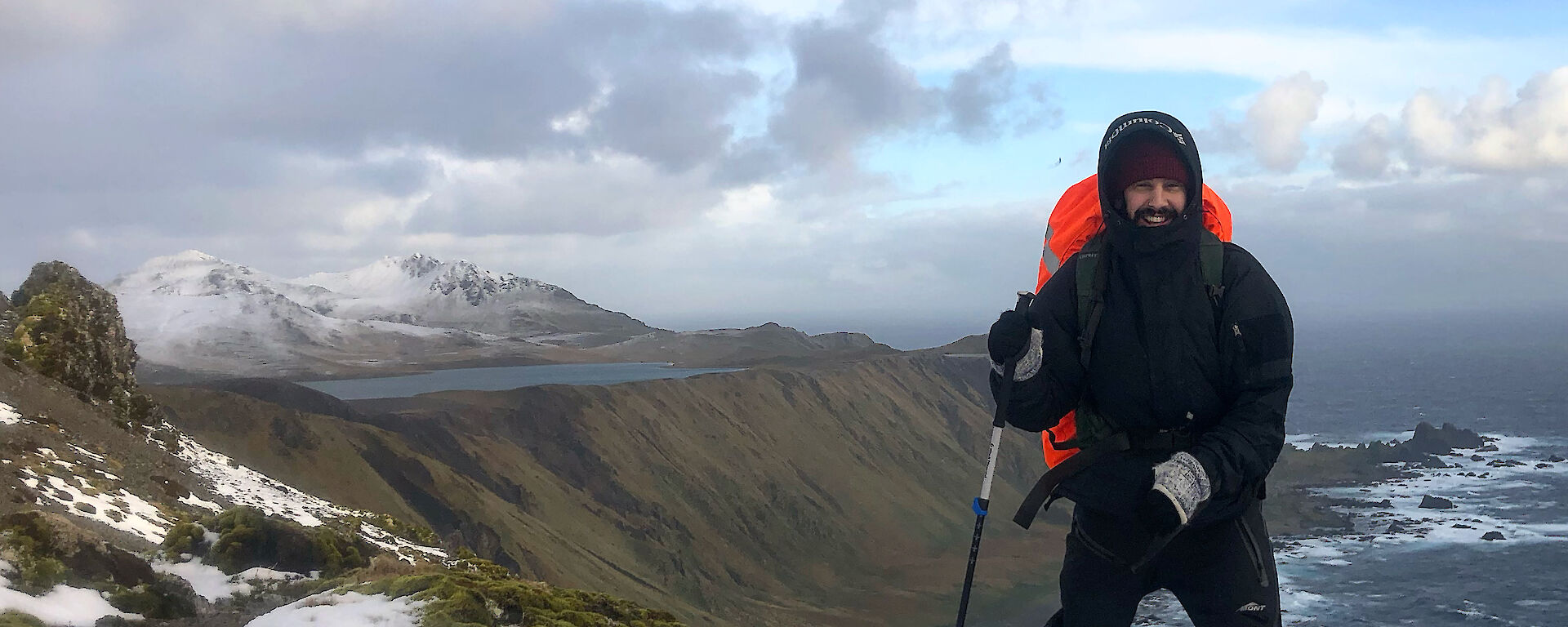 A man in all weather gear and a backpack stands on the snowy ridge of a mountain.  The sea and another snow capped mountain can be seen in the background.