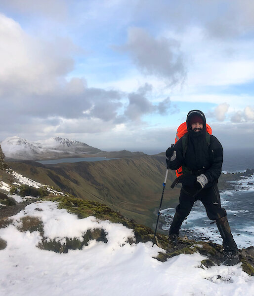 A man in all weather gear and a backpack stands on the snowy ridge of a mountain.  The sea and another snow capped mountain can be seen in the background.
