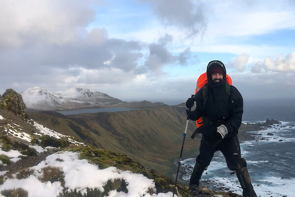 A man in all weather gear and a backpack stands on the snowy ridge of a mountain. The sea and another snow capped mountain can be seen in the background.