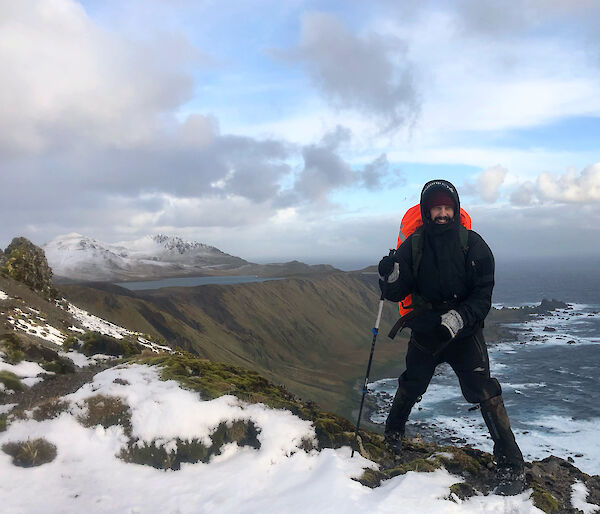 A man in all weather gear and a backpack stands on the snowy ridge of a mountain.  The sea and another snow capped mountain can be seen in the background.