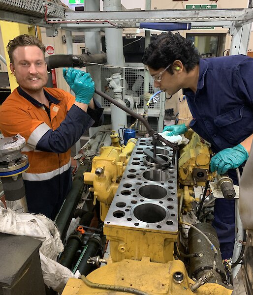 2 men in a workshop repairing the inside of a generator engine.  One is smiling to camera, the other examining the parts.