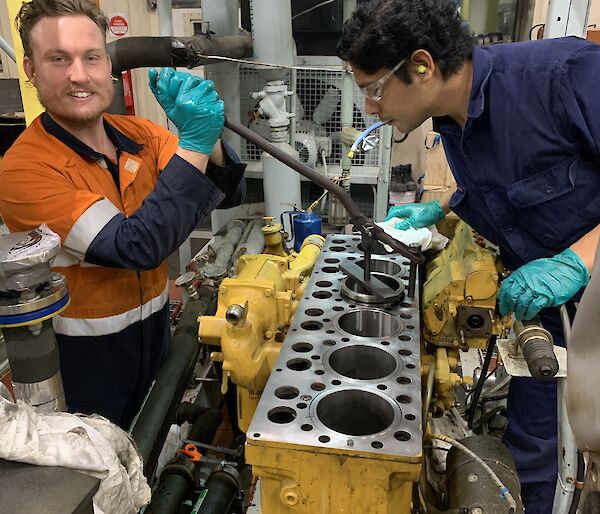 2 men in a workshop repairing the inside of a generator engine. One is smiling to camera, the other examining the parts.