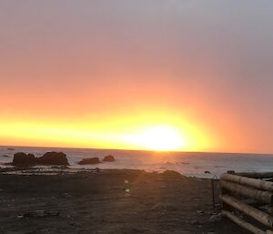 An orange and yellow setting sun with the sea shore in the foreground and a wooden fenced off area to the right