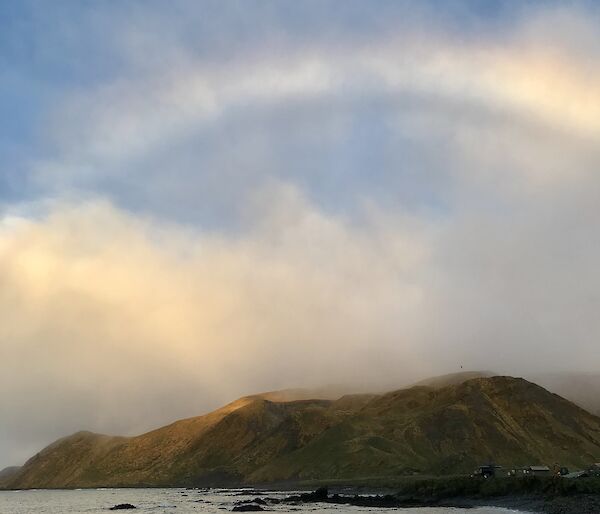 A rainbow in the sky above a green hill, with waves on the shore in the foreground