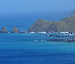 A view from a hill across a blue sea, with the green hilly island shore visible to the right of shot, together with a series of small rock formations