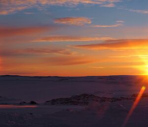 A landscape image with the foreground silhouetted against an blue and orange sky.  A sun dog can be seen just above the horizon.