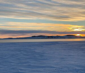 A large expanse of sea ice with a landform on the horizon, against a sky lit by the midday sun.