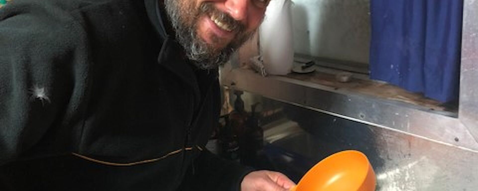 A man smiling to camera with a tin of fruit pudding on the bench in front of him.  He holds a spatula and a bowl in his hands.