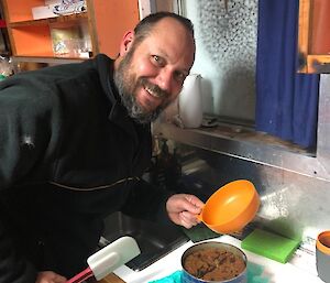 A man smiling to camera with a tin of fruit pudding on the bench in front of him.  He holds a spatula and a bowl in his hands.