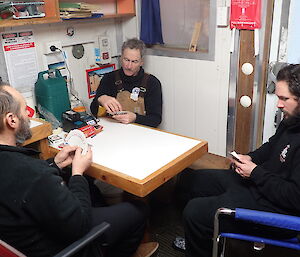 3 men sitting at a small table playing cards in a field hut