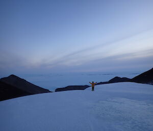 Expeditioner in his yellow deep field jacket standing with arms up in the air on a large blizz tail at Mt Henderson.