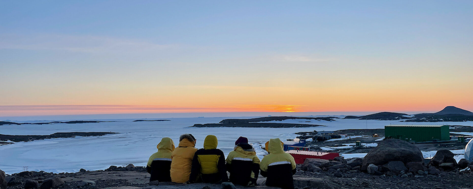 Five expeditioner sitting on a large rock as they watch the sun go down which produces a nice orange glow on the horizon.