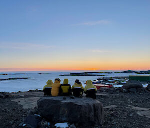 Five expeditioner sitting on a large rock as they watch the sun go down which produces a nice orange glow on the horizon.