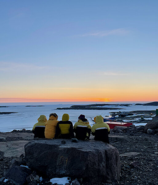 Five expeditioner sitting on a large rock as they watch the sun go down which produces a nice orange glow on the horizon.
