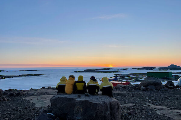 Five expeditioner sitting on a large rock as they watch the sun go down which produces a nice orange glow on the horizon.
