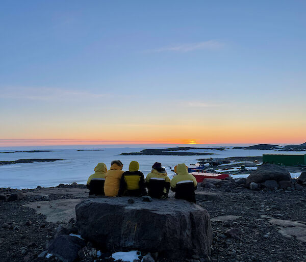 Five expeditioner sitting on a large rock as they watch the sun go down which produces a nice orange glow on the horizon.