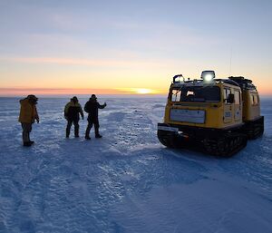 Three expeditioner standing next to the yellow Hagglund over snow vehicle with the orange glow of the sun setting in the background.