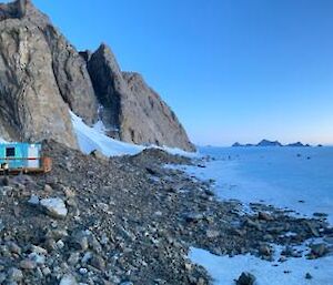 The blue Rumdoodle hut at the base of the rocky cliffs with the yellow Hagglund over snow vehicle in the foreground.