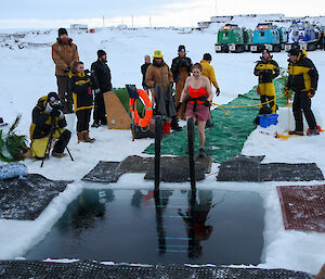 A woman with a rope attached around her waiste approaches a square hole cut in the sea ice. Other expeditioners look on. Three coloured hagglund vehicles are in the background.