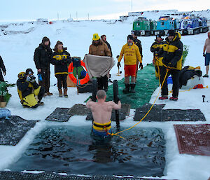 A man with a rope around his waist climbs out of a square pool cut in the sea ice. Another expeditioner waits with a towel, while a group look on.