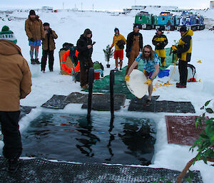 A man carrying a surf board prepares to jump in to a pool of water cut out of sea ice. A small group of expeditioners look on.
