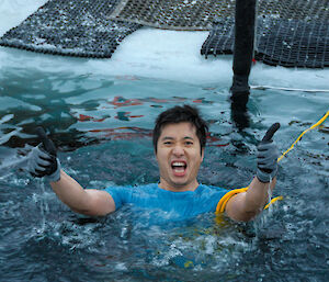 A man wearing black gloves and a blue shirt gives a thumbs up as he floats in a pool of icy water.