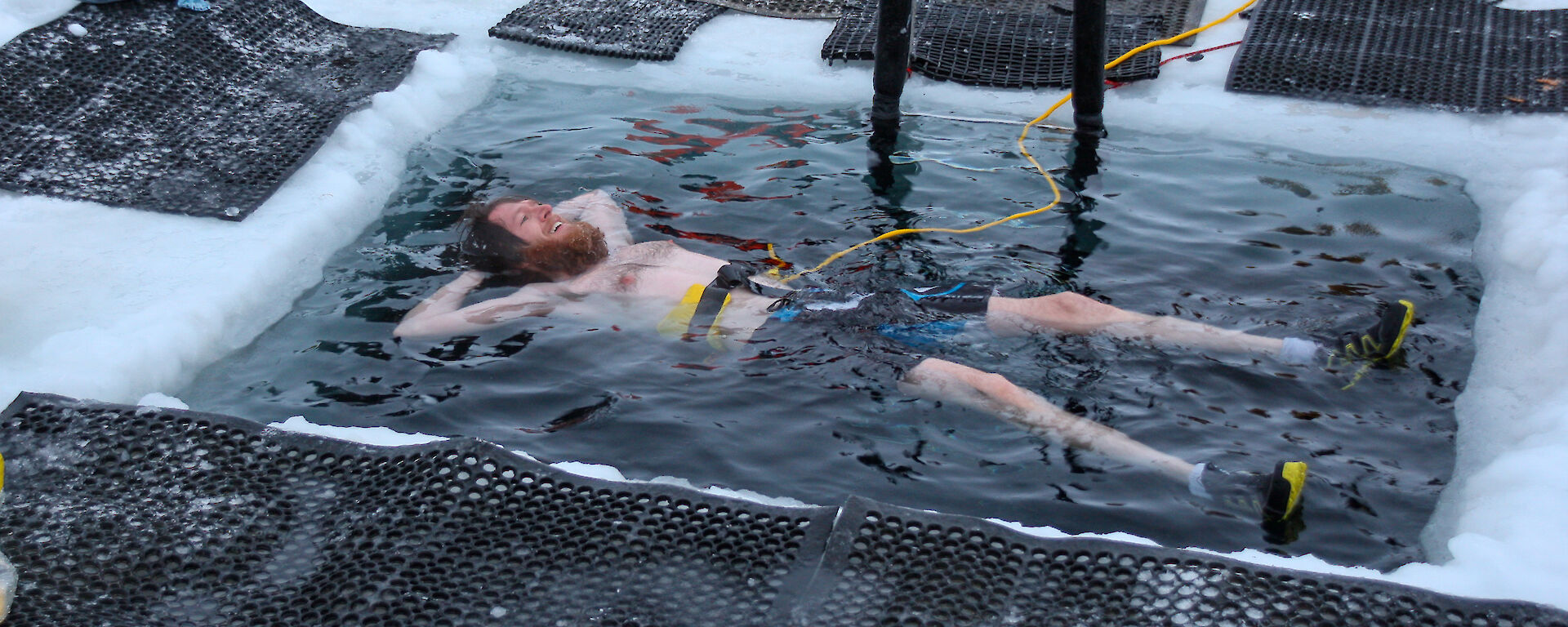 A man floats on his back in a square pool of icy water cut in to the sea ice.