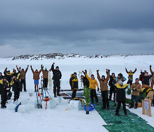 A group of expeditioners raise their arms in triumph as they stand around a pool of water cut in to sea ice.